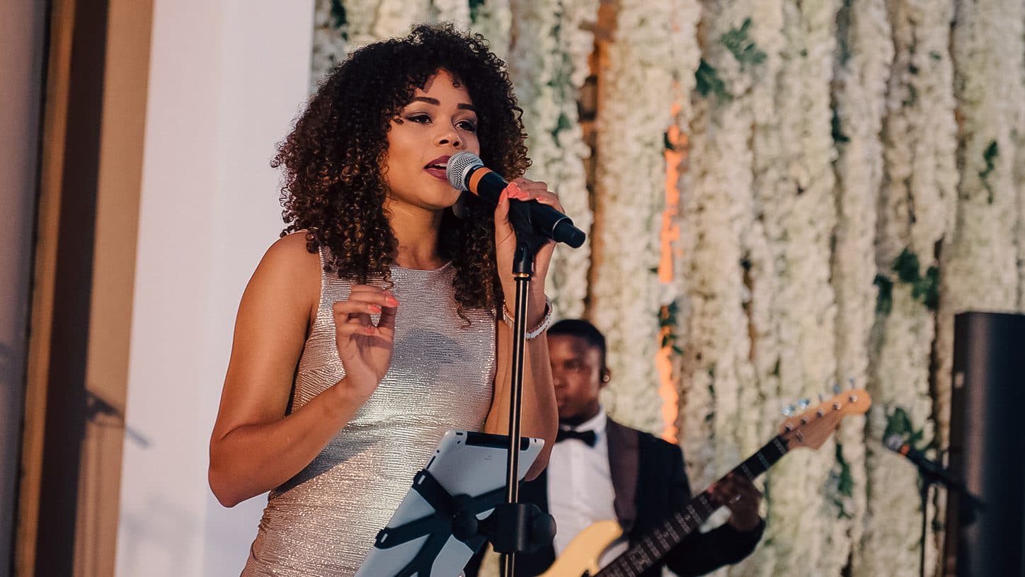 Chanteuse en robe pailletée au micro devant un mur de fleurs blanches, guitariste en retrait
